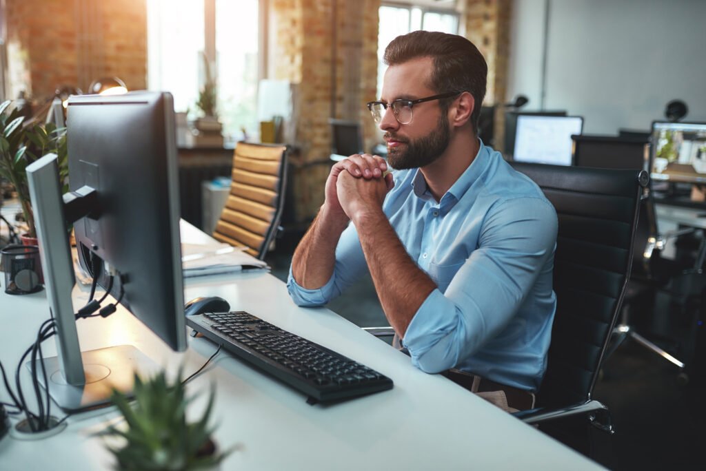 Thinking. Side view of young bearded man in eyeglasses and formal wear working on computer and thinking about branding related questions.